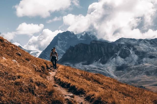 Hiker walking a steep dry-grass mountain trail below rugged peaks